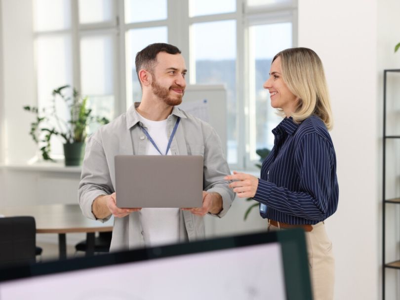 A software engineer holds a laptop while discussing a project with a fellow engineer in an office