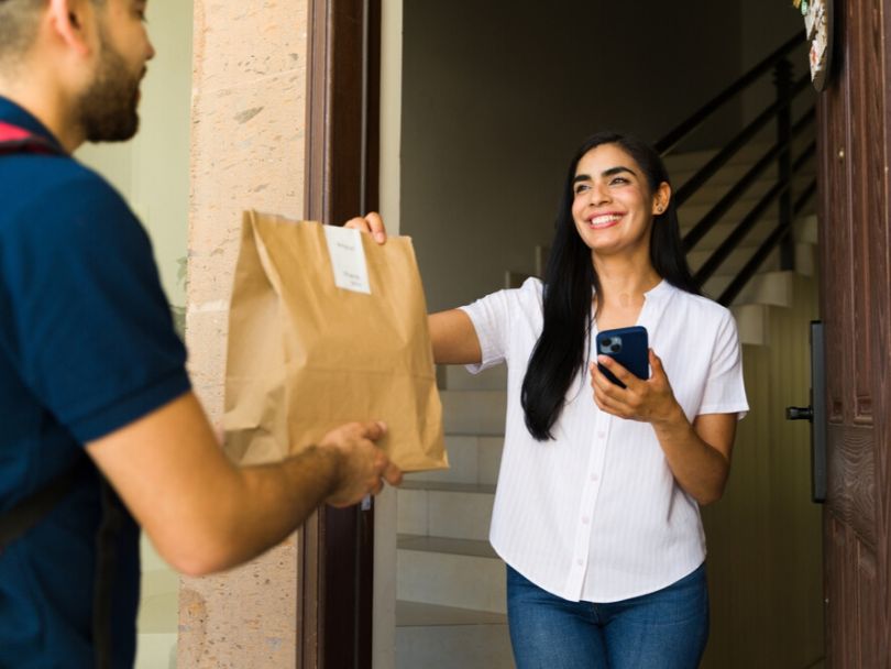 A food delivery courier hands a smiling woman her food order at her home’s front door