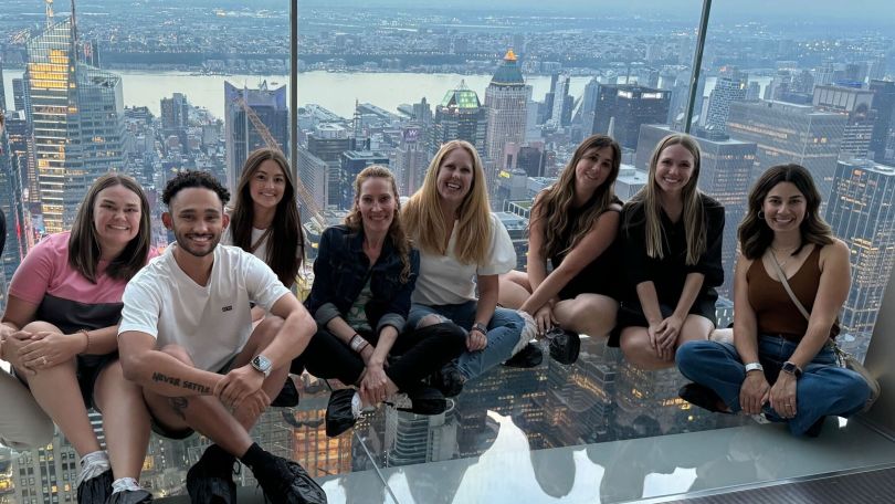 A group of Huntress employees sit on the glass observation floor in a skyscrapper.