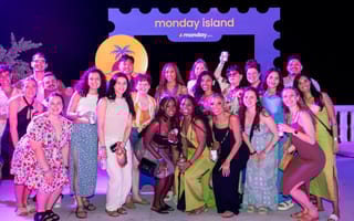 A group of monday employees pose outside by a sign that says "monday island" at night on a beach scene.