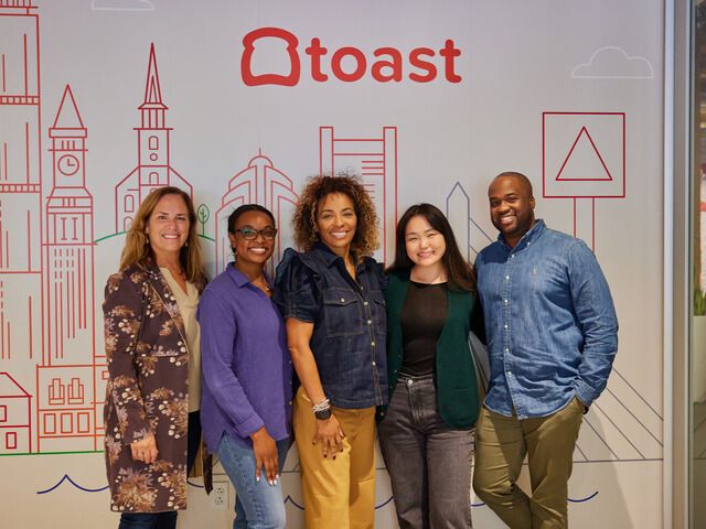 Toast team members posing for a group photo in front of a wall emblazoned with the company’s logo inside one of the company’s offices