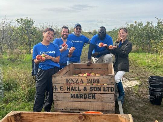 Toast team members in company-branded t-shirts posing for a group photo at an apple orchard while holding apples in their hands